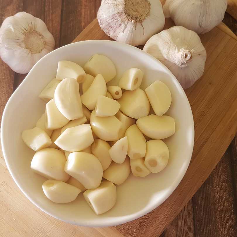 White bowl filled with chopped garlic cloves on a wooden surface with whole garlic bulbs.