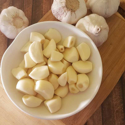White bowl filled with chopped garlic cloves on a wooden surface with whole garlic bulbs.